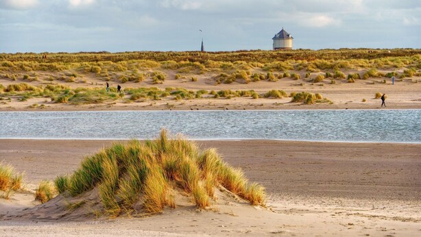 Una playa con dunas de arena, una masa de agua y una estructura distante.