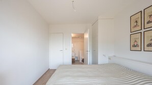 A bedroom with a striped bedspread, a wooden floor, white walls, and a door leading to a bathroom.