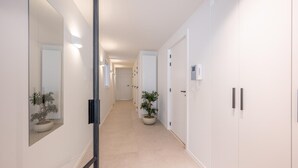 Un couloir aux murs blancs, avec un plancher de couleur claire et une plante en pot. Il y a des portes sur le côté droit et un grand miroir sur le côté gauche.