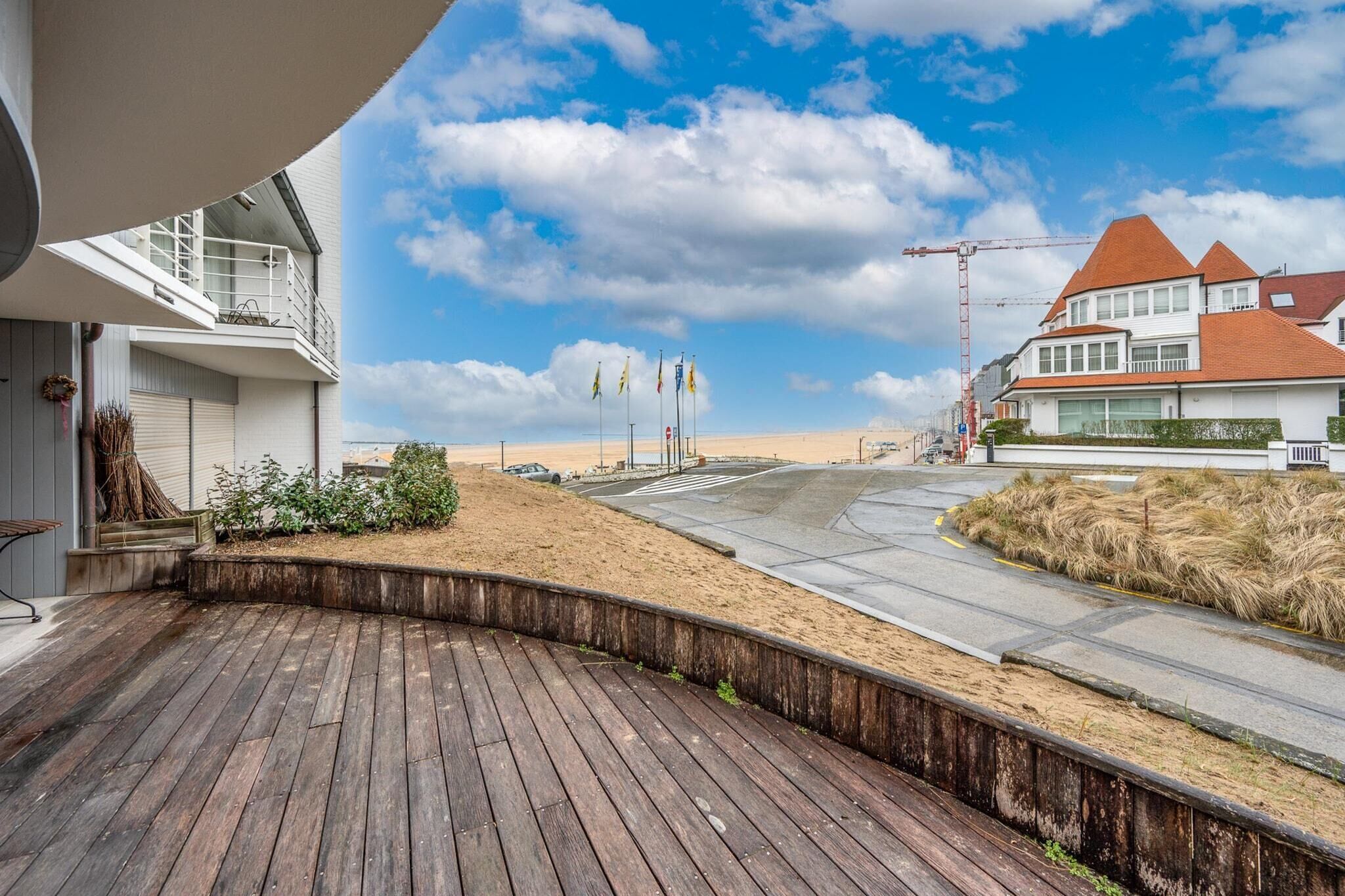 A wooden deck with a curved edge, a sandy area with grass, and a road with a construction crane in the background.