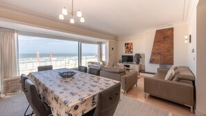 Uma sala de estar ampla com uma janela grande de frente para a praia, uma mesa de jantar com toalha de mesa estampada, um sofá e uma televisão.