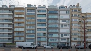 Um edifício residencial de vários andares com varandas, uma carrinha branca estacionada em frente e vários carros ao longo da rua.