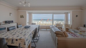 Ampla sala de estar com uma grande janela com vista para a praia, área de jantar com mesa e um espaço de estar aconchegante.