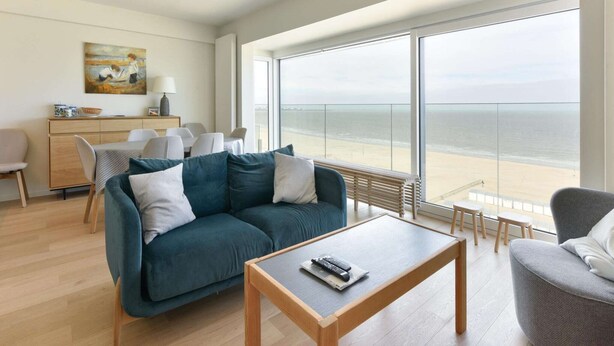 Un salon moderne doté d’une grande fenêtre donnant sur la plage, d’un canapé, d’une table basse et d’un coin repas avec des chaises.