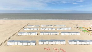 Une plage bordée de rangées de cabines blanches, modulaires et facilement transportables, et une zone délimitée par une clôture où se trouve du matériel de plage aux couleurs vives.
