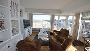Amplia sala de estar con un gran ventanal que ofrece vistas de la ciudad. Hay un mueble blanco para entretenimiento, un conjunto de sofás marrones y una mesa de centro.