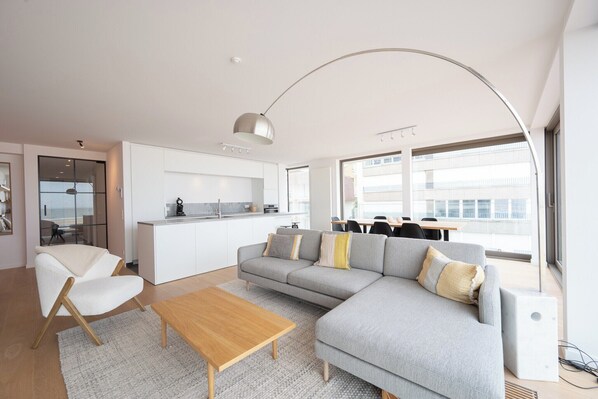 A modern living room with a grey sofa, a wooden coffee table, and a white armchair. A kitchen with white cabinets and a large window is visible in the background.