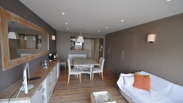 Een moderne hotelkamer met een houten vloer, een witte bank, een eettafel met stoelen, een keukenhoek en een grote spiegel.