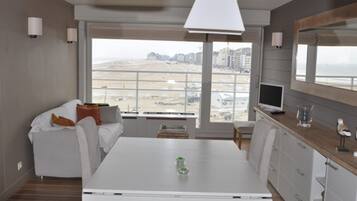 Een moderne woonkamer met een witte bank, een eettafel en een groot raam met uitzicht op een strand en gebouwen.