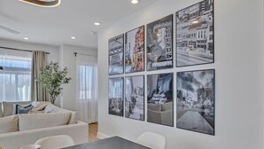 Una sala de estar moderna con un sofá, una mesa de comedor y un ventanal grande. Varias fotografías enmarcadas decoran la pared.