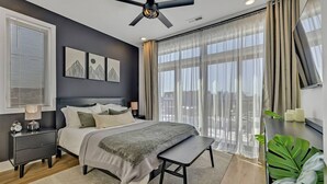 Un dormitorio moderno con una cama, mesitas de noche, un ventilador de techo y un amplio ventanal con cortinas.