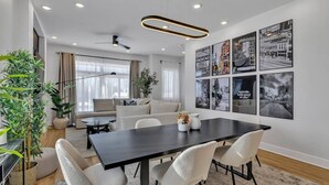 Una sala de estar moderna con una mesa de comedor de madera oscura, sillas blancas y un sofá. Hay varias imágenes enmarcadas en la pared, un ventilador de techo y un ventanal con cortinas.