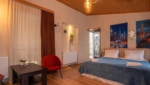 A hotel room with a bed, a desk, a chair, a radiator, a window with curtains, and a cityscape painting. - Hotel and Restaurant VILLA GIVISI (Lagodekhi)