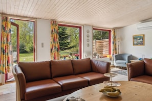 A living room with a brown leather sofa, a wooden coffee table, and a view of a garden through large windows. - Charming holiday home in peaceful surroundings (Gedser)