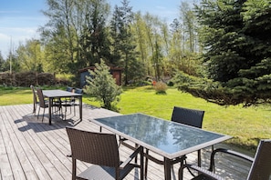 A wooden deck with a glass-top table and chairs, surrounded by a grassy yard and trees. - Charming holiday home in peaceful surroundings (Gedser)