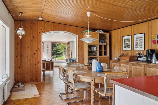 Une pièce aux murs lambrissés, comprenant une table à manger, des chaises, un coin cuisine et une vue sur l’extérieur, visible à travers une porte en arc de cercle.