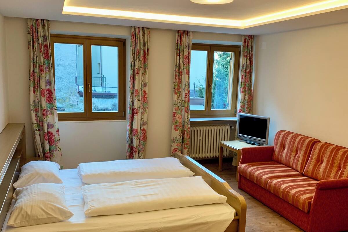 A hotel room with a bed, a red sofa, a TV, and windows with floral curtains.