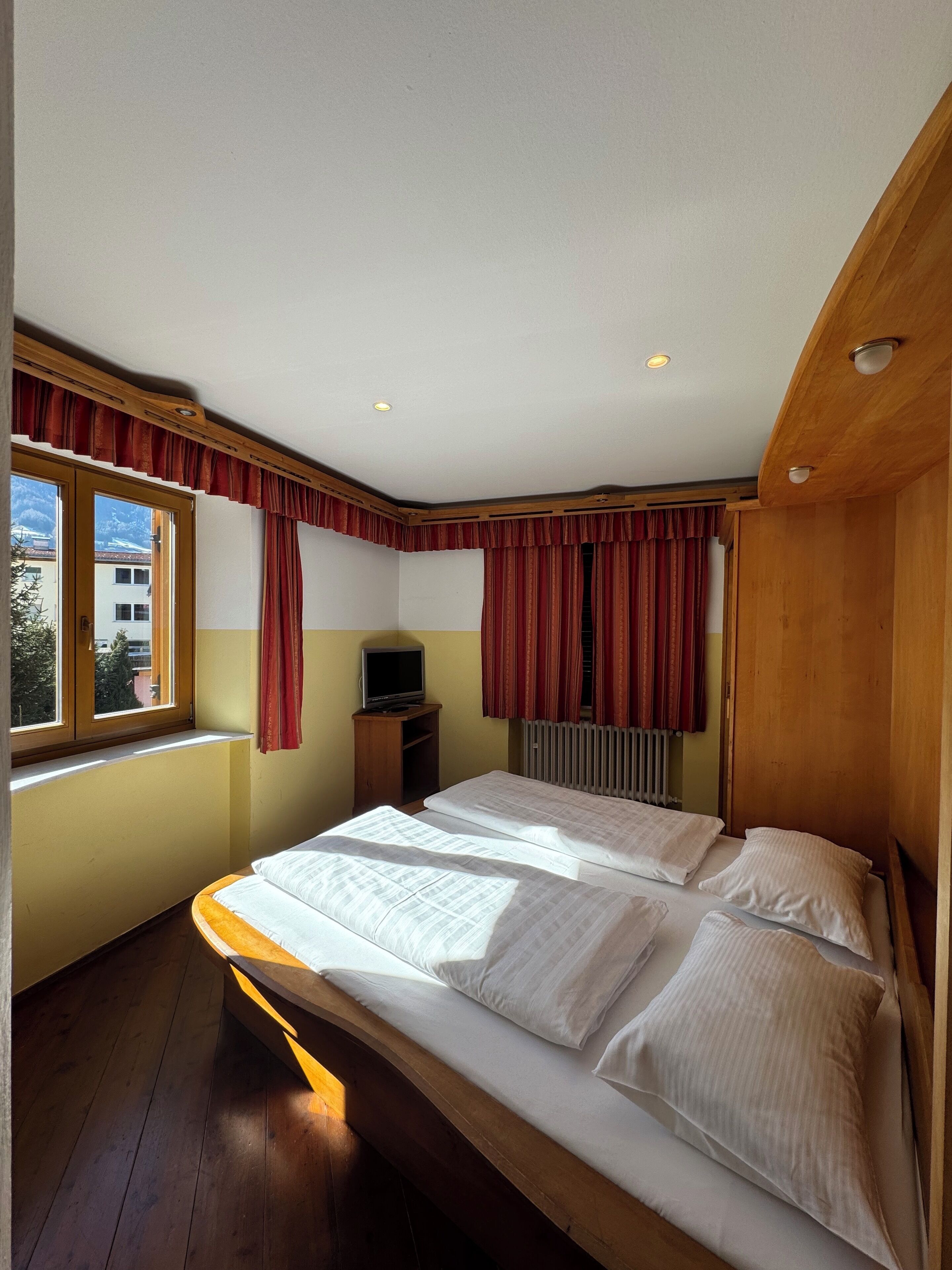 A bedroom with a wooden bed, a television on a small stand, and a window with curtains.
