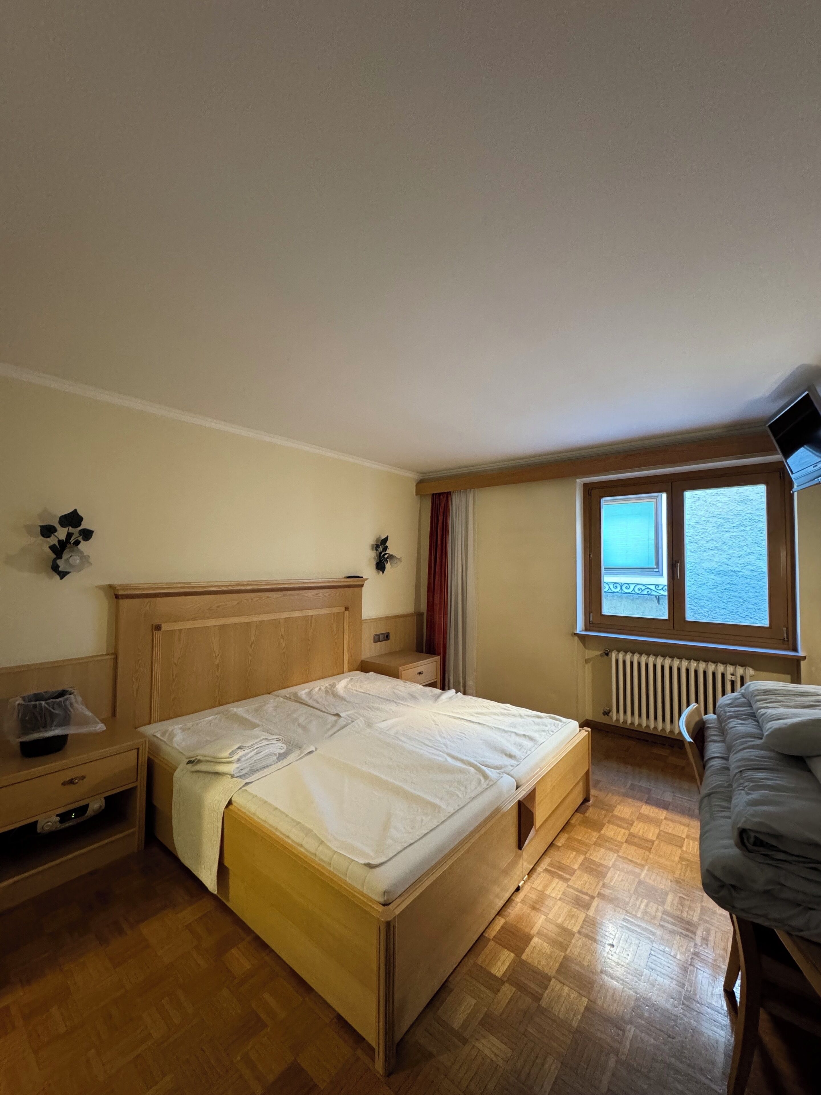 Camera da letto con un letto e un comodino in legno, una cassettiera, una finestra vista e un termosifone.