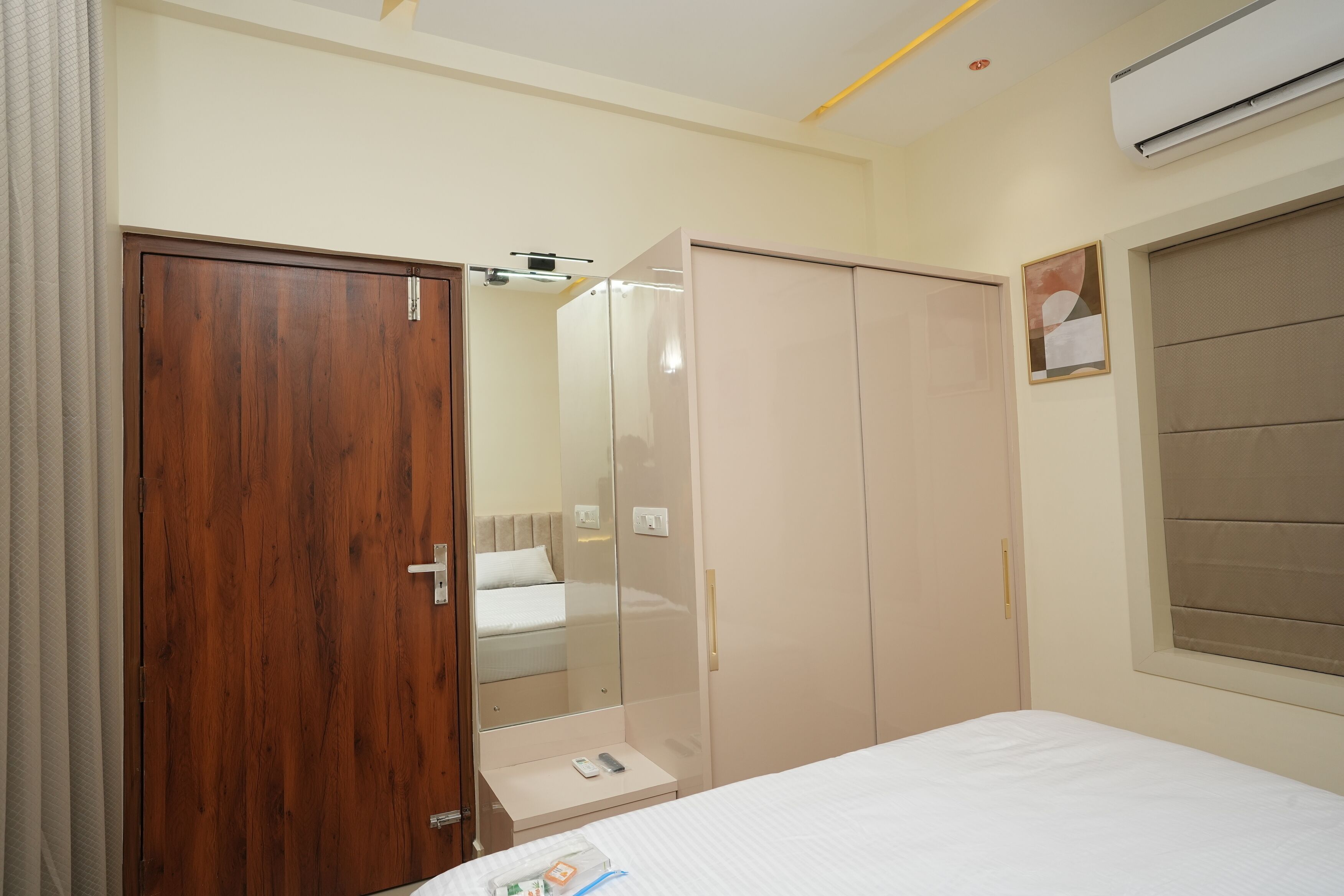 Un dormitorio con un armario de madera, un armario con espejo, una mesita de noche con un teléfono y un libro, y una cama con edredón blanco.