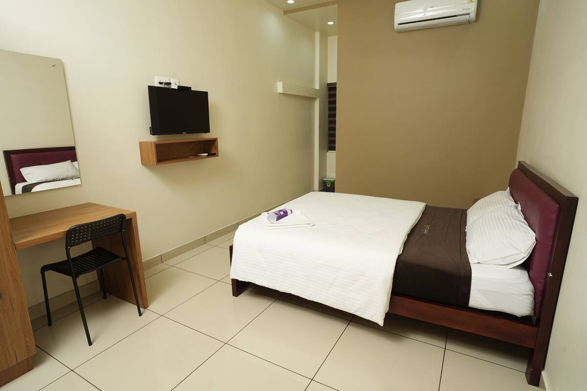 A hotel room with a bed, a desk, a chair, a TV, and an air conditioner.
