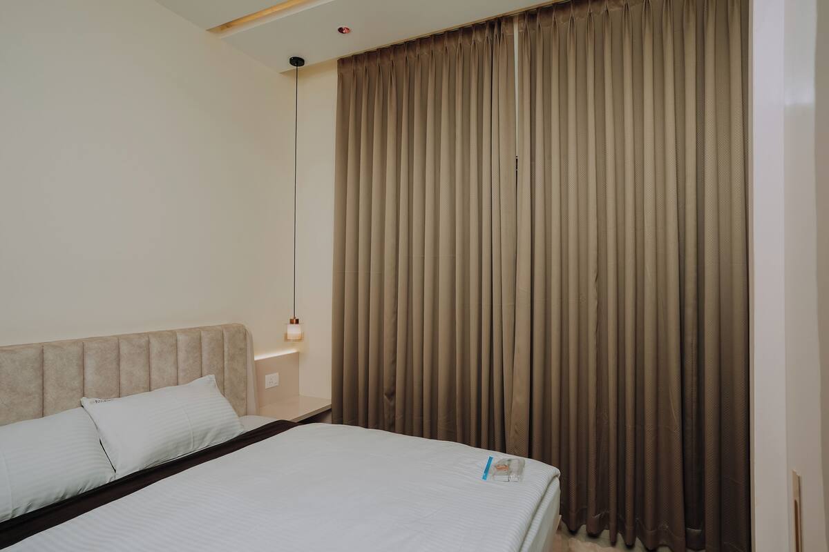 A hotel room with a bed, a headboard, a wall-mounted light, and a window with curtains.