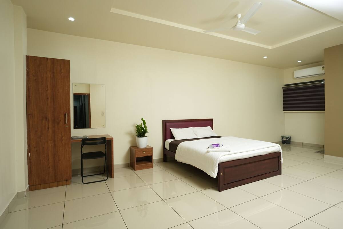 A bedroom with a bed, a desk, a chair, a wardrobe, a ceiling fan, and an air conditioning unit.