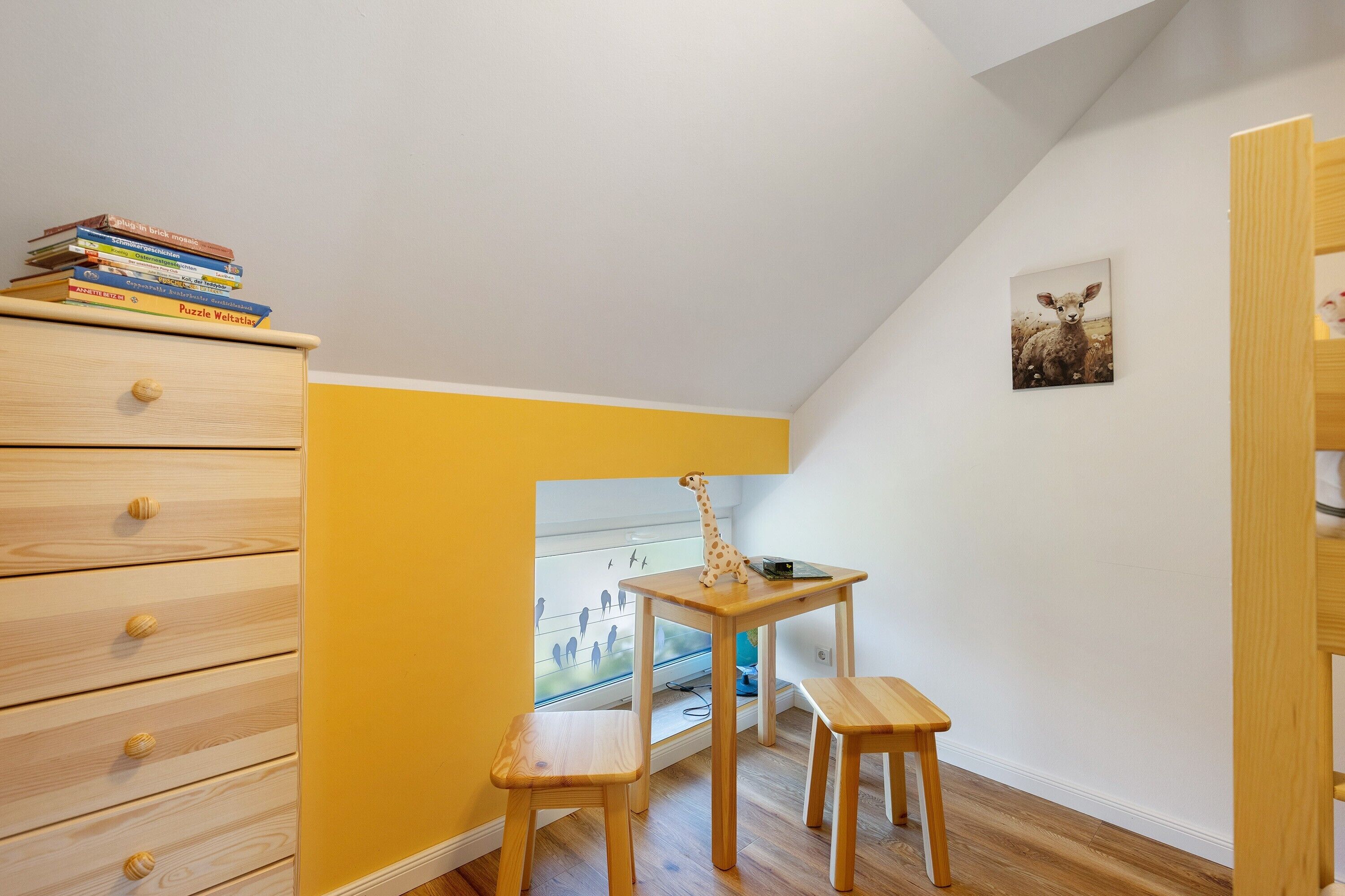 Quarto de criança com cômoda de madeira, uma pequena escrivaninha com um brinquedo de girafa e um quadro com a imagem de uma ovelha.