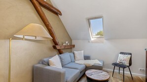 A loft bedroom with a sloped ceiling, a sofa, a chair, a lamp, a small table, and a window.