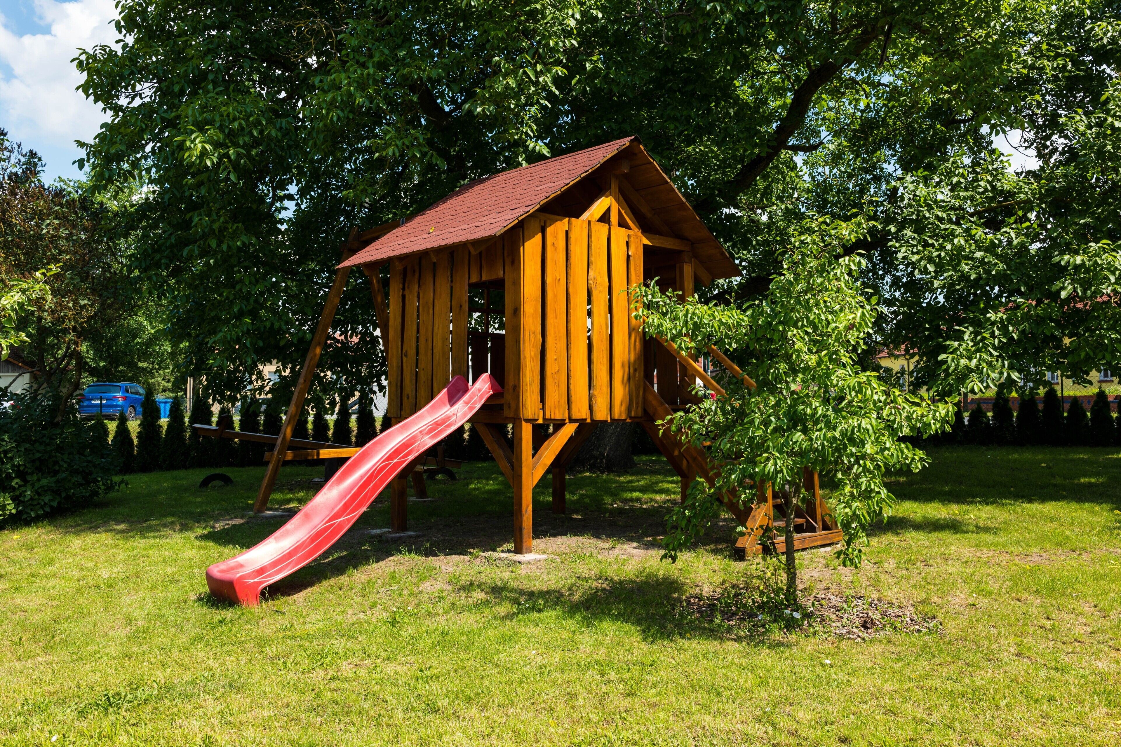 A wooden playhouse with a red slide in a grassy area with trees.