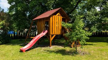 A wooden playhouse with a red slide in a grassy area with trees.