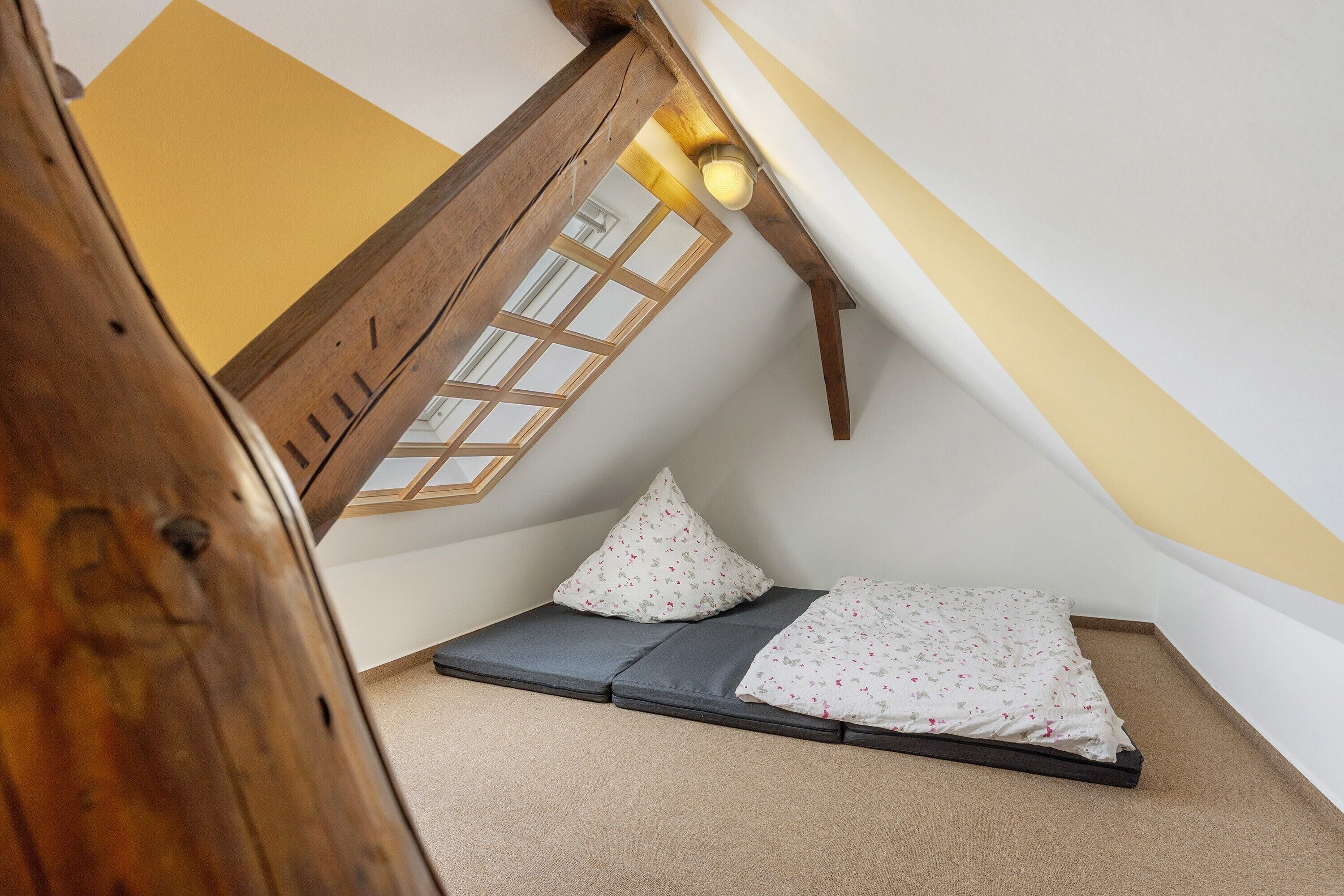 A small, cozy attic room with a sloped ceiling, a window with wooden lattice, and a bed with a patterned blanket.