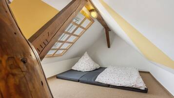 A small, cozy attic room with a sloped ceiling, a window with wooden lattice, and a bed with a patterned blanket.