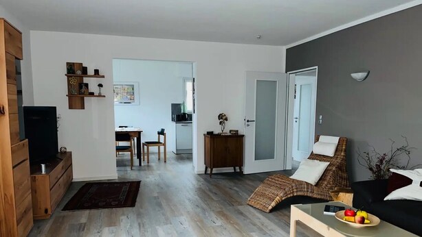 Ein modernes Wohnzimmer mit einer Couch, einem Couchtisch und einem Fernseher. Es gibt einen Essbereich mit Tisch und Stühlen, und im Hintergrund eine Küche.