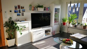 Una sala de estar moderna con un televisor de pantalla plana, muebles blancos, una mesa de centro de vidrio y diversas plantas en macetas.