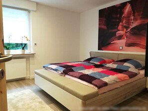 Un dormitorio con cama, ventana con plantas, radiador y un cuadro en la pared.