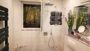 A modern bathroom with a shower, a toilet, a sink with toiletries, a mirror, and a window with a forest view. - Apartment 'Am Silberg' with Mountain View, Private Terrace, and Wi-Fi (Plettenberg)