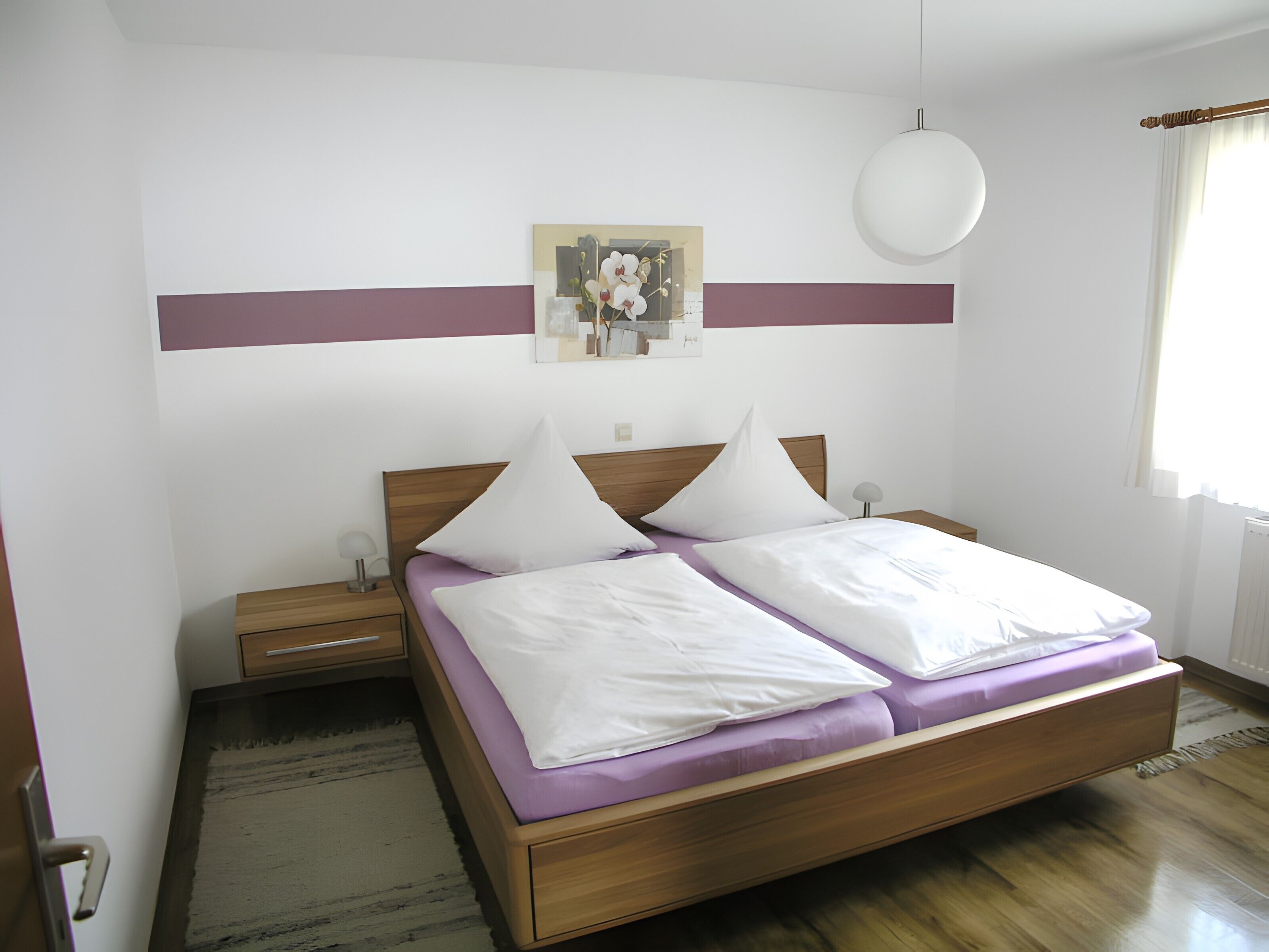 A bedroom with a wooden bed, white pillows, a nightstand, a wall-mounted lamp, and a painting.
