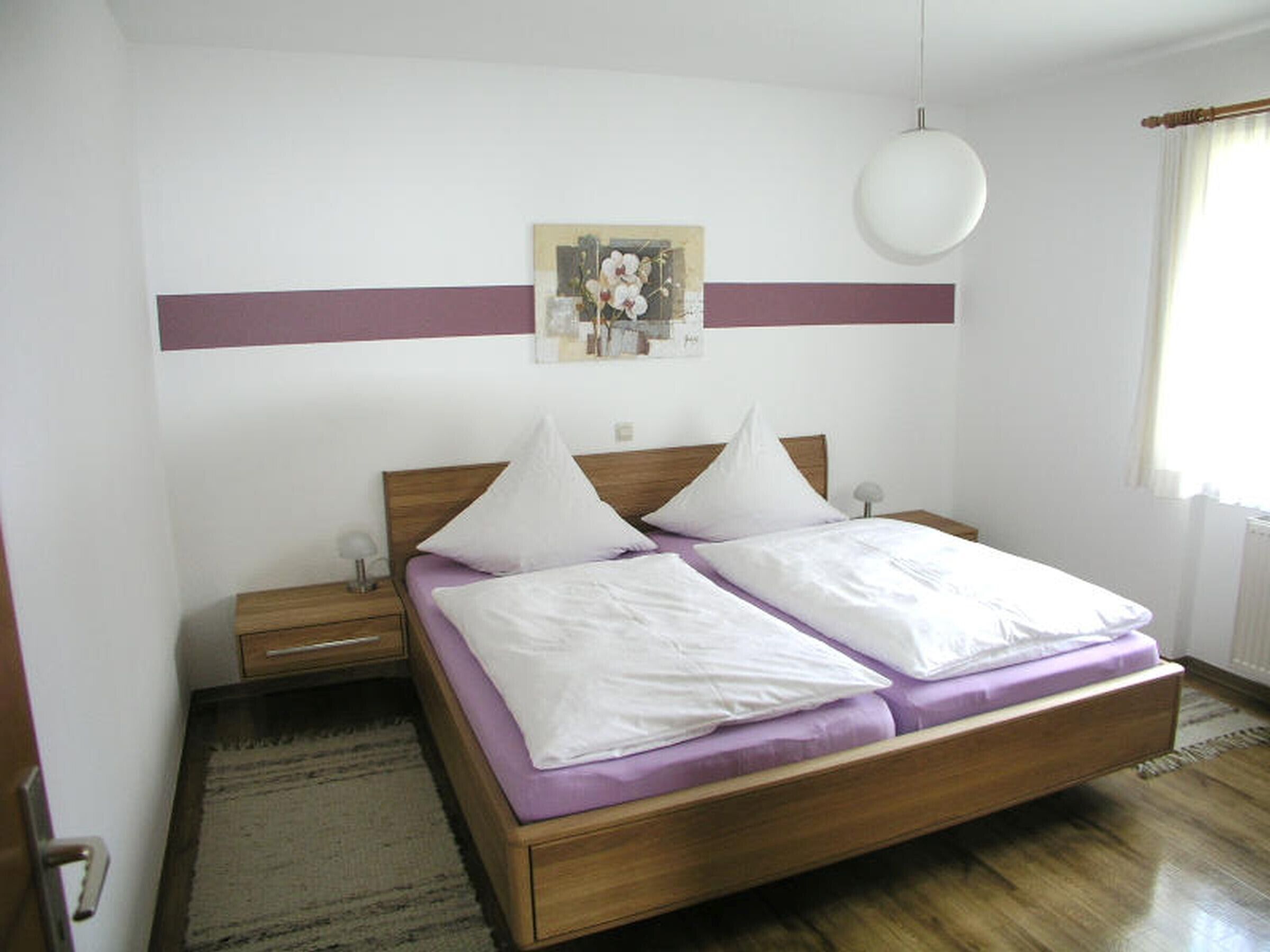 Ein Schlafzimmer mit einem hölzernen Bett, zwei Nachttischen, einem Wandleuchter, einem Gemälde und einem Fenster mit Vorhängen.