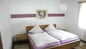 Une chambre à coucher avec un lit en bois, deux tables de chevet, une commode, une lampe fixée au mur, un tableau et une fenêtre avec des rideaux.