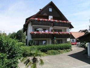 En bygning med flere etager med balkoner pyntet med blomster, en belagt indkørsel og en palme i forgrunden.