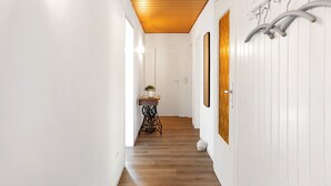 Um corredor com piso de madeira, paredes brancas, teto com painéis de madeira, uma porta, uma pequena mesa com um vaso e um porta-roupas fixado na parede.