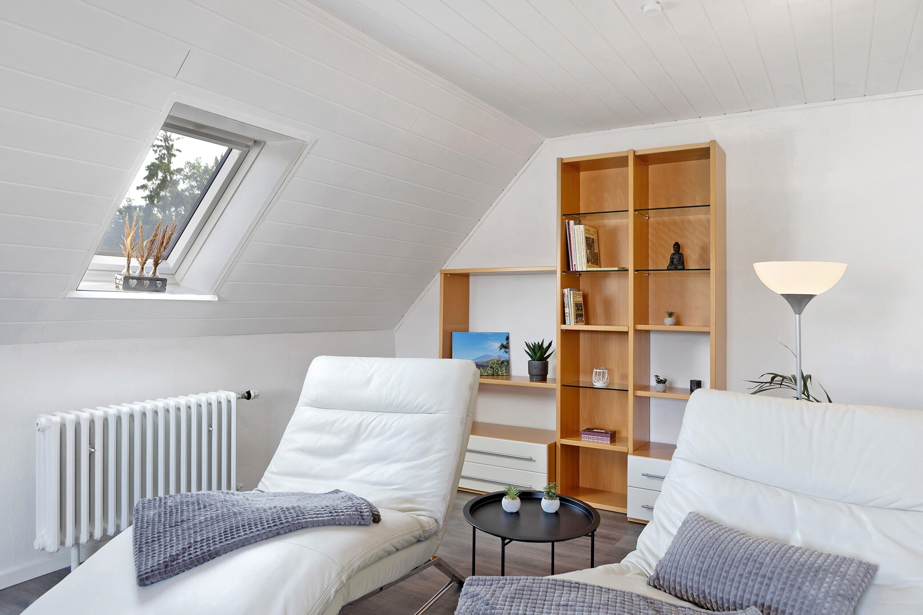 Une chambre de style loft moderne, avec un plafond en pente, des murs blancs, une bibliothèque en bois, un radiateur et une lucarne.