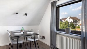 Uma sala de jantar com uma mesa, cadeiras, um vaso de planta e uma janela com vista para casas.