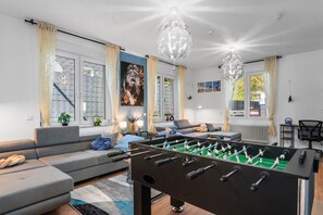 Una sala de estar moderna con mesa de fútbol, sofás grises, comedor y ventanas grandes con cortinas.