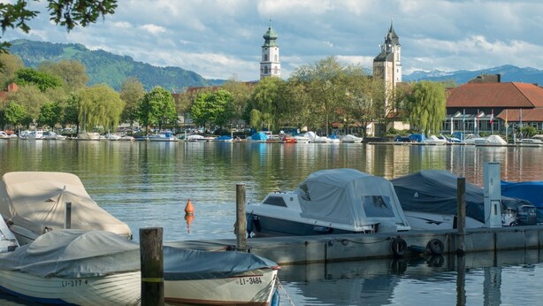 A marina with boats docked, a church with a tower, and a mountain backdrop.