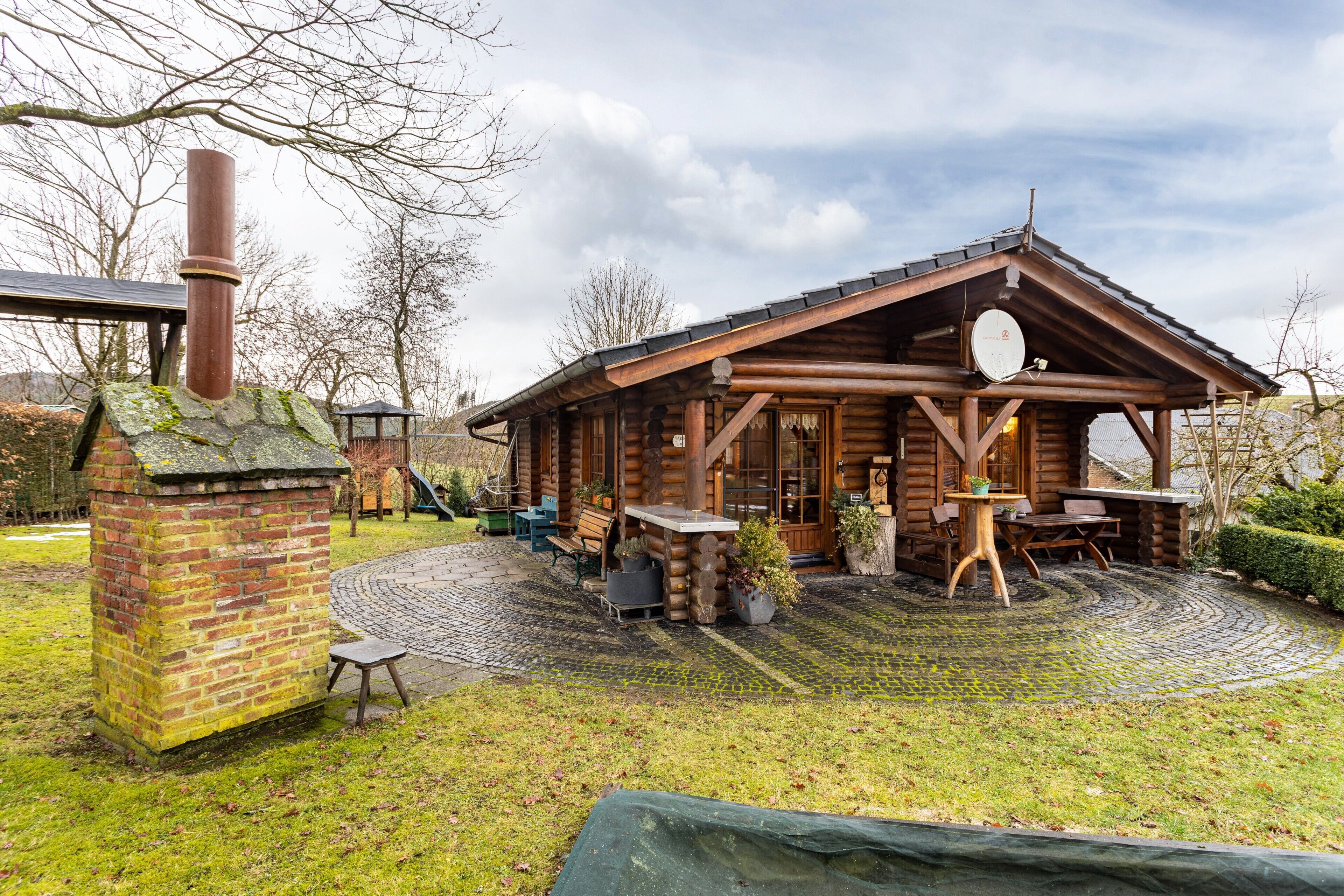 A rustic log cabin with a chimney, outdoor seating, and a brick structure.