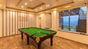 A pool table in a room with striped wallpaper, a large window with a view, and a ceiling with recessed lighting. - The Hosteller Kufri, Shimla (Theog)