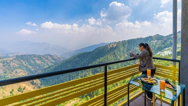 A balcony with a clear view of a mountainous landscape, a glass table with food and drinks, and a person standing by the railing. - The Hosteller Kufri, Shimla (Theog)
