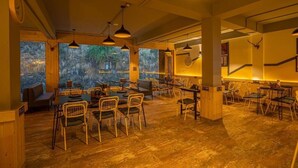 A restaurant with wooden furniture, pendant lighting, and large windows offering a view of the outdoors. - The Hosteller Kufri, Shimla (Theog)